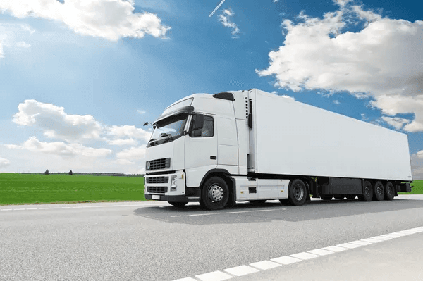 White semi-truck driving on a highway with a grassy field and blue sky in the background.